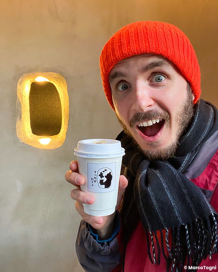 Marco Togni sorridente tiene un bicchiere di carta davanti al Kuma no Te Cafe, Osaka, dove una zampa di orso serve i clienti.
