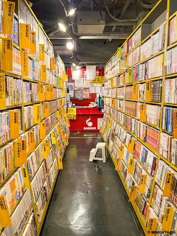 Corridoio interno del negozio Mandarake con scaffali gialli pieni di manga e un bancone rosso sul fondo.