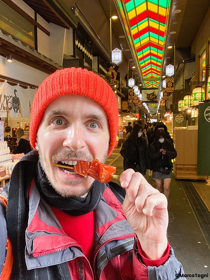 Marco Togni con berretto rosso mangia spiedini di pesce crudo tra le bancarelle del mercato di Nishiki a Kyoto.