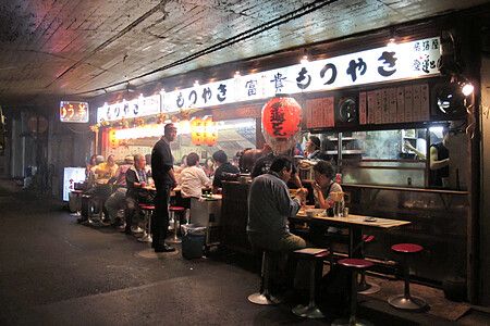 Persone sedute in una izakaya sotto un viadotto ferroviario a Yurakucho, in Giappone, circondate da insegne luminose.