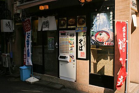 La facciata di un piccolo ristorante a Tokyo con un distributore automatico di ticket e insegne rosse.