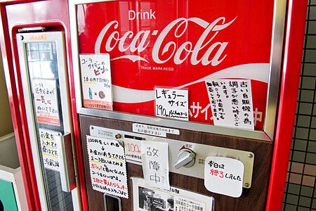 Un distributore automatico di bevande Coca-Cola rosso con numerosi avvisi scritti a mano in giapponese attaccati.