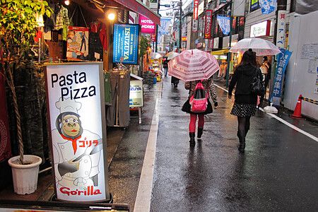 Strada piovosa a Shimokitazawa, Giappone, con insegna "Pasta Pizza" e persone con ombrelli.