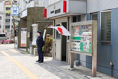 Un agente di polizia in divisa è in piedi davanti all'ingresso di un koban su un marciapiede urbano.