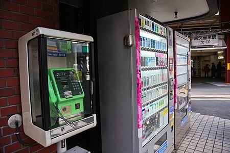 Una cabina telefonica verde e distributori automatici di sigarette vicino all'ingresso nord della stazione Higashikurume.