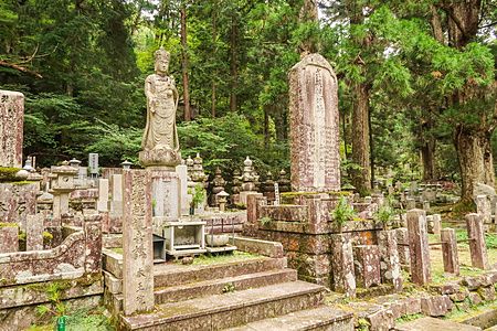 Tombe e scultura al cimitero di Okunoin, Monte Koya, tra alberi verdi.