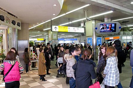 Un gruppo di persone staziona all'interno della stazione ferroviaria di Shinjuku a Tokyo sotto una segnaletica gialla.