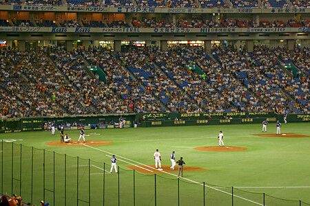 Una partita di baseball giocata sul campo verde dello stadio Tokyo Dome davanti a un numeroso pubblico sugli spalti.