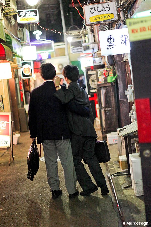 Due persone in abito formale camminano abbracciate di spalle in un vicolo notturno a Golden Gai, Shinjuku.