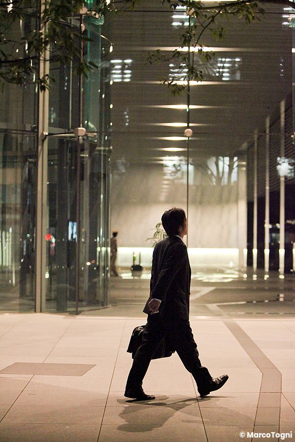 Un uomo in abito scuro cammina di profilo durante una camminata serale salaryman in Giappone, davanti a una vetrata.