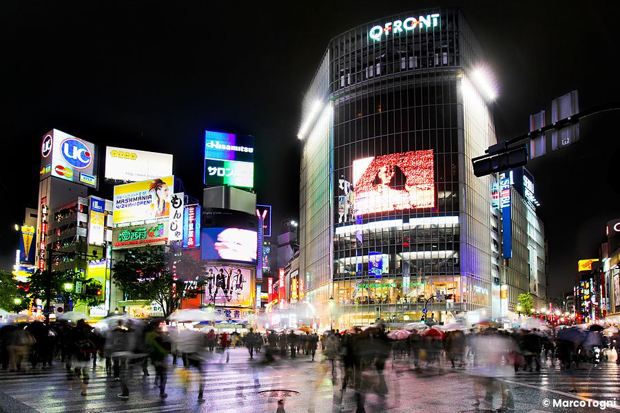 Incrocio di Shibuya a Tokyo con pioggia e persone sotto ombrelli.