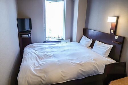 Letto matrimoniale in una camera d'albergo compatta con pareti color crema e una finestra luminosa.