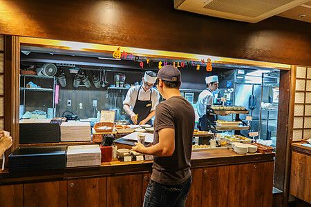 Un uomo al bancone di una cucina aperta mentre i cuochi preparano colazione.