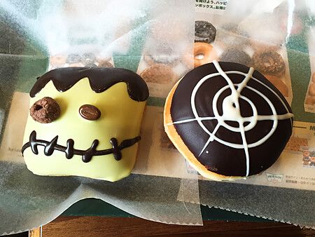 Due ciambelle decorate a tema Halloween su carta cerata, una a forma di mostro verde e una con ragnatela al cioccolato.
