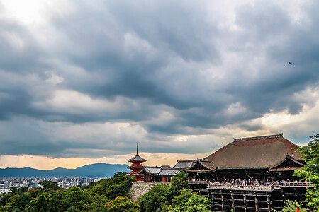 Veduta panoramica del tempio Kiyomizudera a Kyoto con una pagoda rossa e persone sulla terrazza sotto un cielo nuvoloso.
