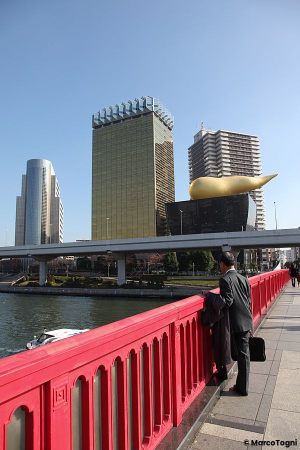 Un uomo in giacca e cravatta osserva l'Asahi Beer Hall e lo skyline dal ponte rosso sul fiume Sumida a Tokyo.