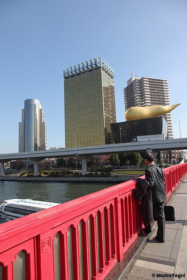 Uomo in giacca guarda l'Asahi Beer Hall di Tokyo da un ponte rosso.