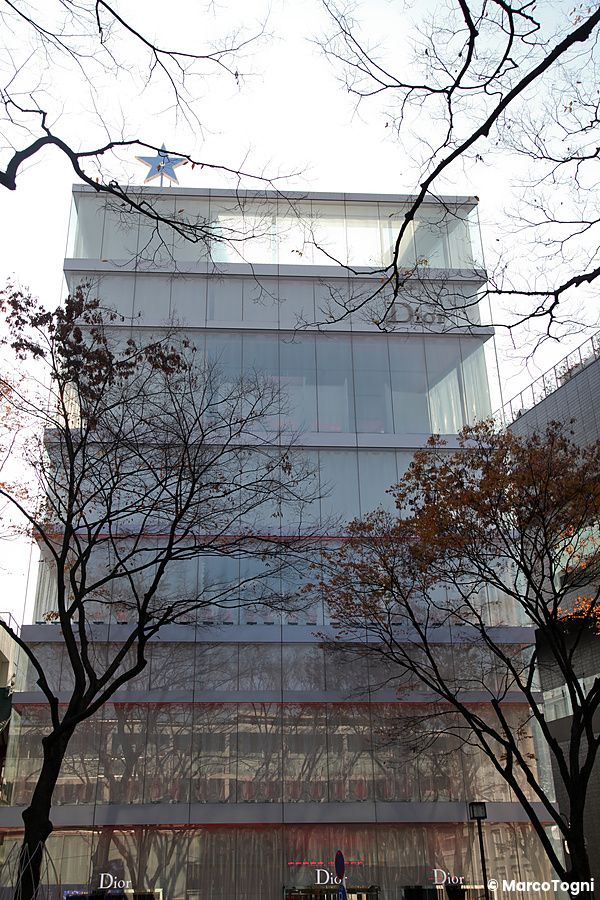 Edificio di vetro a Omotesando, Tokyo, con alberi spogli attorno.