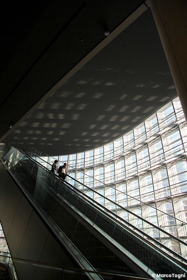 Persone su scala mobile in un edificio moderno con tetto ondulato.