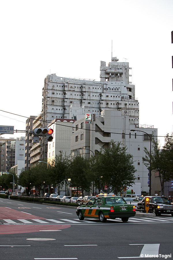 Incrocio a Shinjuku con il New Sky Building sullo sfondo e un taxi di passaggio.