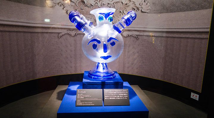 Un'anfora in vetro veneziano con decorazioni a forma di volto esposta su un piedistallo blu al Hakone Glass Forest Museum.