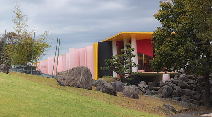 Edificio moderno dai colori accesi inserito in un parco con rocce, alberi e un prato inclinato in pendenza.