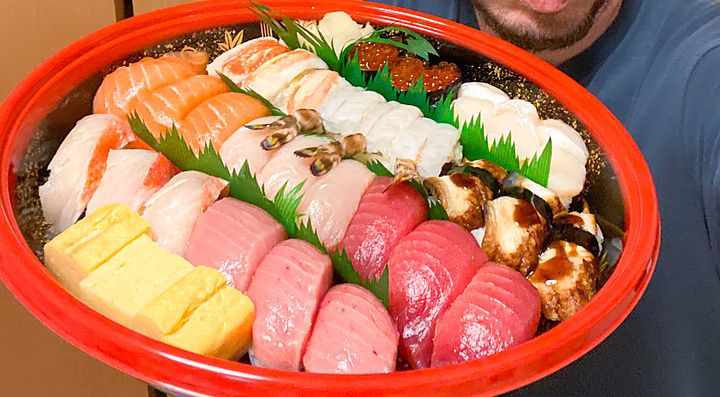 Marco Togni regge un grande vassoio di plastica rossa pieno di vari tipi di sushi nigiri davanti a una parete.