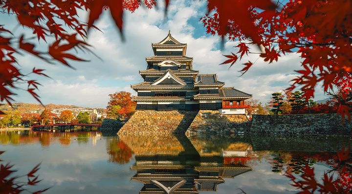 Veduta del castello di Matsumoto incorniciato da foglie di acero rosso autunnali con il suo riflesso nell'acqua.