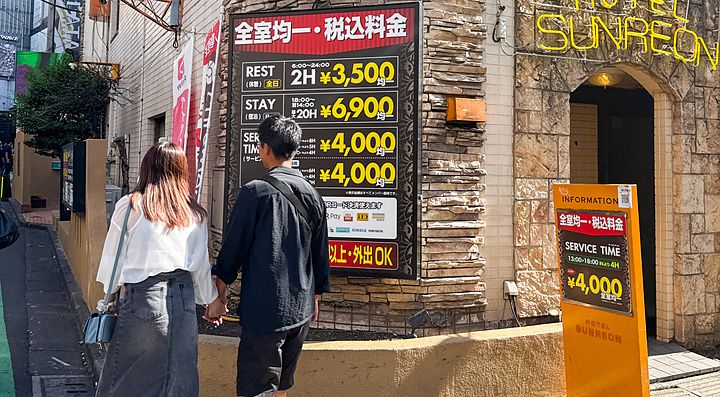 Una coppia cammina mano nella mano davanti all'Hotel Sunreon a Shibuya sotto la luce del giorno.