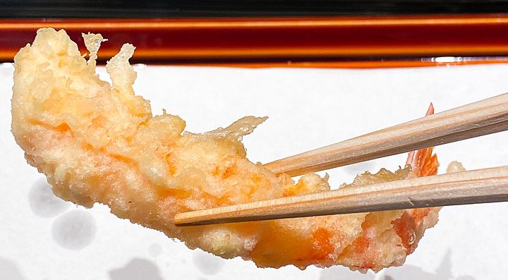 Un gambero a Tempura Kondo sollevato da un paio di bacchette di legno sopra un vassoio coperto di carta.