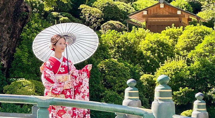 Una donna in kimono rosso tiene un ombrello bianco su un ponte in un giardino a Tokyo.