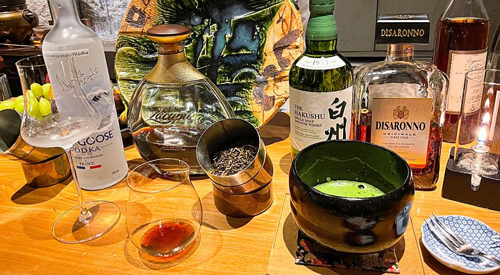 Bottiglie di alcolici, una ciotola di matcha e un piatto decorativo sul bancone in legno del Mixology Salon a Ginza.