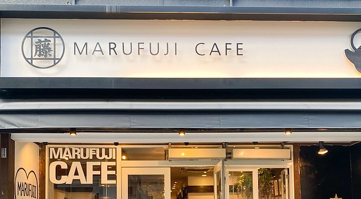L'ingresso a Marufuji, un caffè con insegna bianca, vetrate, albero di Natale all'esterno e persone all'interno.