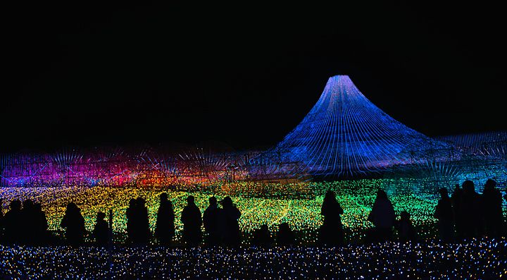Sagome di visitatori che osservano una grande installazione luminosa a forma di monte Fuji durante un festival notturno.