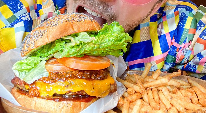 Marco Togni con occhiali rosa morde un hamburger in un ristorante.