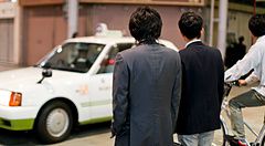 Due persone in abito formale attendono un taxi bianco in una strada urbana in Giappone.