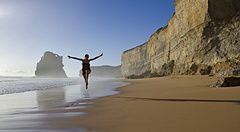 Donna felice che salta sulla spiaggia accanto a scogliere imponenti.
