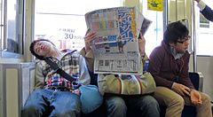 Tre passeggeri siedono su un treno pendolari in Giappone, con uno dei soggetti che dorme e un altro che legge il giornale.