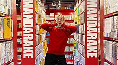 Marco Togni in maglione rosso posa in un corridoio tra scaffali di manga nel negozio Mandarake in Giappone.