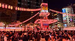 Una folla di persone partecipa a un matsuri ad Ebisu davanti alla stazione, con un palco centrale illuminato da lanterne.