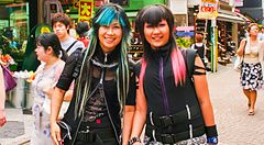 Due persone sorridenti vestite in modo alternativo camminano a Shibuya tra la folla.