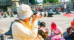 Una persona con cappello chiaro fotografa individui in costume ad Harajuku durante una giornata di sole.
