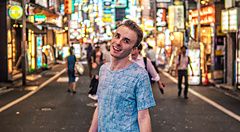 Marco Togni sorridente in primo piano su una strada illuminata dalle insegne al neon nel quartiere di Shinjuku.