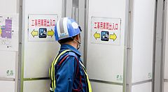 Un operaio giapponese con elmetto davanti a barriere e cartelli.