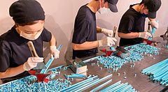 Tre persone con maschere e guanti tagliano lunghi bastoncini di caramelle artigianali azzurre su un tavolo in acciaio.