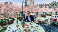 Marco Togni siede a un tavolo su una terrazza a Roppongi, circondato da ciliegi in fiore con un grattacielo sullo sfondo.