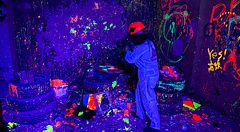 Una persona con tuta e casco distrugge oggetti nella rage room a Reeast Room, in un ambiente illuminato da luci UV.