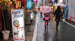 Strada piovosa a Shimokitazawa, Giappone, con insegna "Pasta Pizza" e persone con ombrelli.