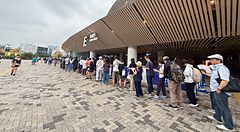Una fila di persone in attesa all'ingresso dello Stadio Nazionale Giapponese a Tokyo su un piazzale pavimentato.