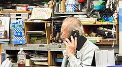 Uomo anziano al telefono in piccolo ufficio giapponese affollato.